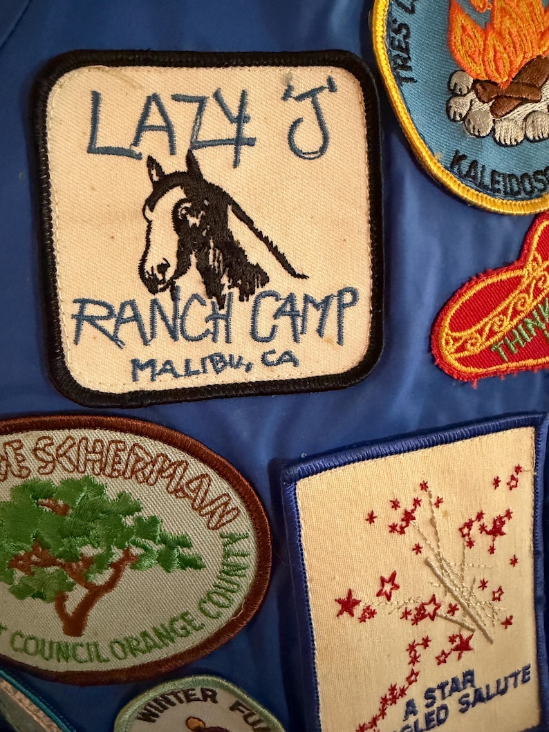 Puede incluir: Colecci&oacute;n de parches bordados sobre fondo azul. Uno de los parches dice "LAZY J RANCH CAMP MALIBU, CA" con una ilustraci&oacute;n de un caballo. Otros parches presentan texto e ilustraciones, incluyendo una fogata, estrellas y un &aacute;rbol.