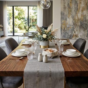 Pode incluir: Uma mesa de jantar de madeira comprida com um caminho de mesa cinzento texturizado, pratos de cer&acirc;mica branca, copos transparentes, talheres dourados e um arranjo floral central num vaso cinzento e dourado.