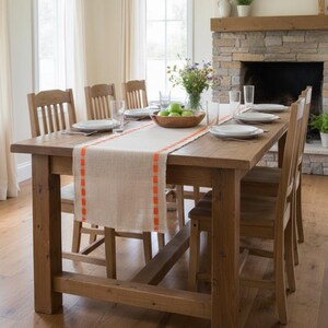 May include: A rectangular wooden dining table with a light-colored burlap table runner featuring bright orange stitched edges. The table is set with white plates, clear drinking glasses, and a wooden bowl filled with green apples in the center.