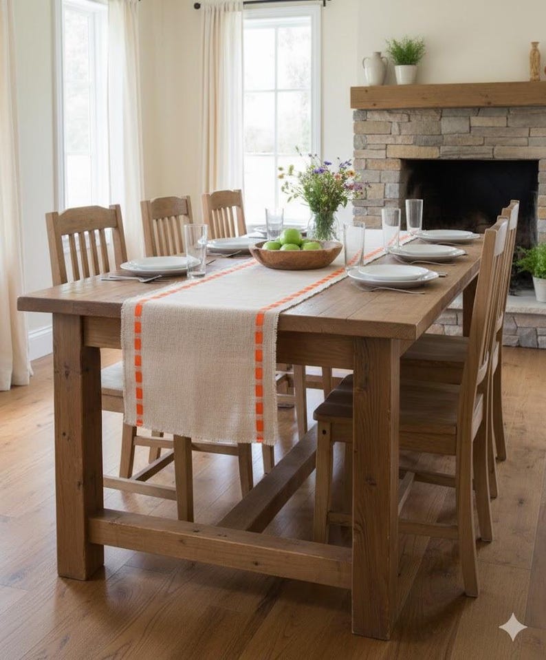 May include: A rectangular wooden dining table with a light-colored burlap table runner featuring bright orange stitched edges. The table is set with white plates, clear drinking glasses, and a wooden bowl filled with green apples in the center.