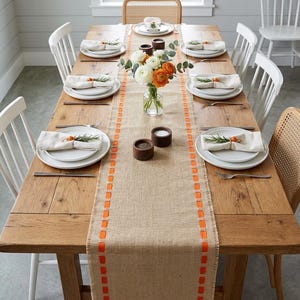 May include: A rectangular wooden dining table set for six with a burlap table runner featuring orange ribbon accents. The table includes white plates, cream-colored napkins with rosemary sprigs, a glass vase with white and orange flowers.