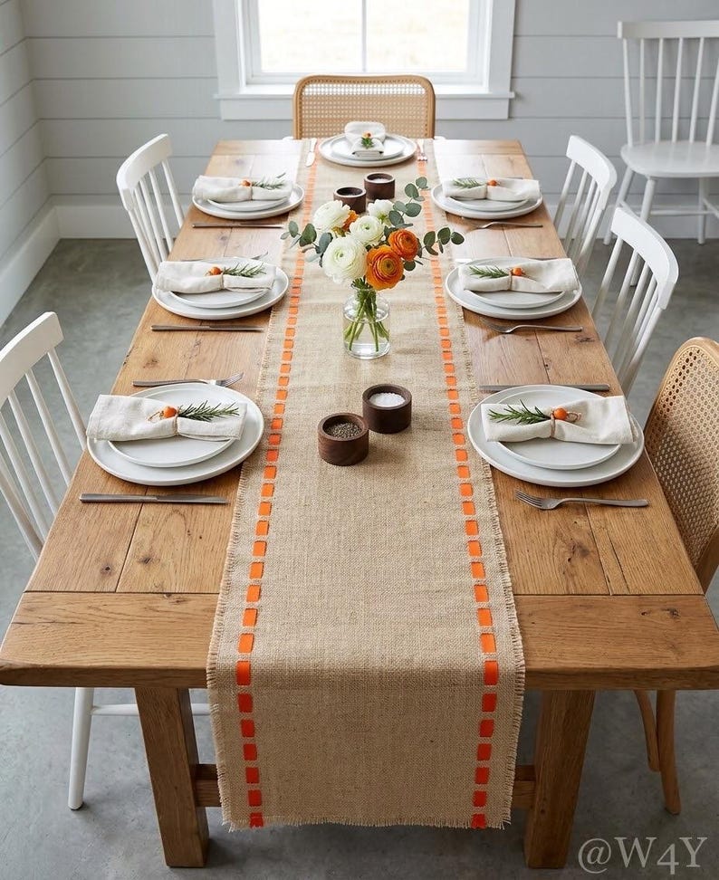 May include: A rectangular wooden dining table set for six with a burlap table runner featuring orange ribbon accents. The table includes white plates, cream-colored napkins with rosemary sprigs, a glass vase with white and orange flowers.