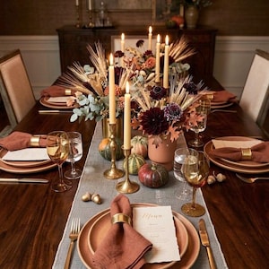 Pode incluir: Uma mesa de jantar com pratos cor de terracota, guardanapos de linho cor de ferrugem com an&eacute;is dourados, talheres dourados e copos &acirc;mbar. O centro de mesa inclui velas compridas em suportes de lat&atilde;o, trigo seco e pequenas ab&oacute;boras.