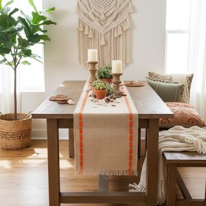 May include: A rectangular beige burlap table runner featuring bright orange stitched vertical borders and frayed edges. The runner is centered on a wooden dining table, decorated with two wooden candle holders, white pillar candles, and small potted succulents.