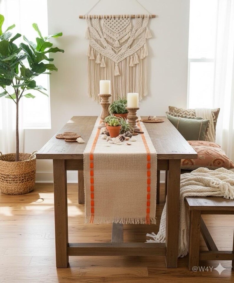 May include: A rectangular beige burlap table runner featuring bright orange stitched vertical borders and frayed edges. The runner is centered on a wooden dining table, decorated with two wooden candle holders, white pillar candles, and small potted succulents.