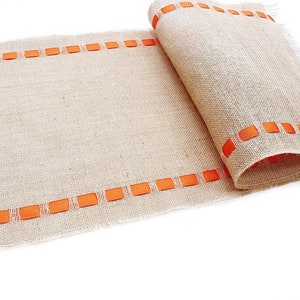 May include: A tan burlap table runner featuring a coarse, woven texture and frayed edges. Two parallel rows of bright orange ribbon are woven through the fabric along the length of the runner, creating a repeating rectangular pattern.