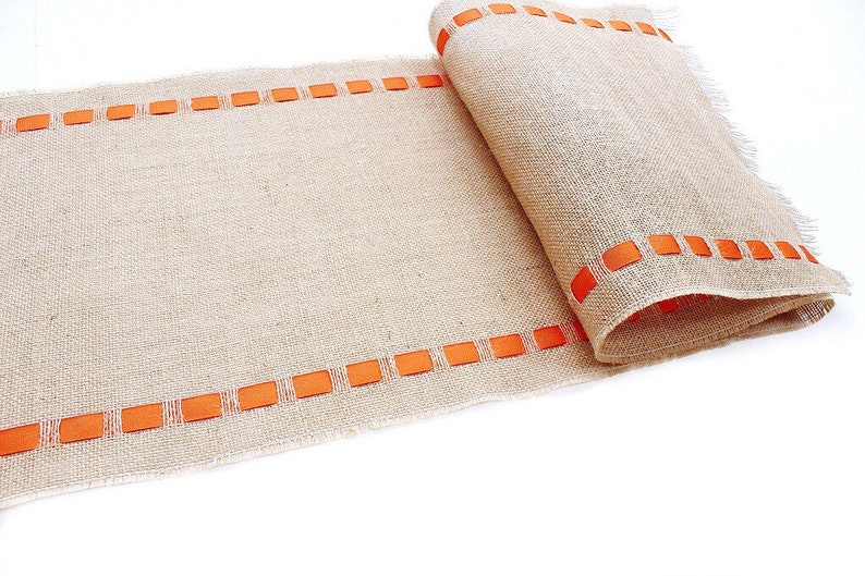 May include: A tan burlap table runner featuring a coarse, woven texture and frayed edges. Two parallel rows of bright orange ribbon are woven through the fabric along the length of the runner, creating a repeating rectangular pattern.