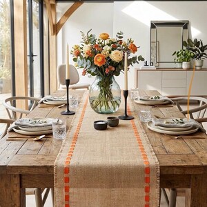May include: A rustic wooden dining table set for a meal, featuring a burlap table runner with orange stitched accents, a clear glass vase filled with orange and cream flowers, two black taper candle holders, and white ceramic plates with napkins.