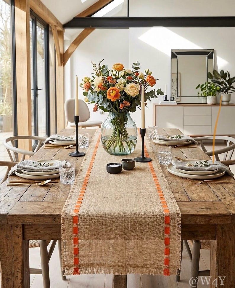 May include: A rustic wooden dining table set for a meal, featuring a burlap table runner with orange stitched accents, a clear glass vase filled with orange and cream flowers, two black taper candle holders, and white ceramic plates with napkins.