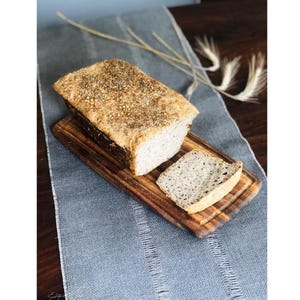 Pode incluir: Um p&atilde;o artesanal retangular com uma crosta dourada coberta de sementes e ervas, sobre uma t&aacute;bua de corte de madeira. Uma fatia est&aacute; cortada e colocada ao lado do p&atilde;o, revelando um miolo interior claro e poroso.