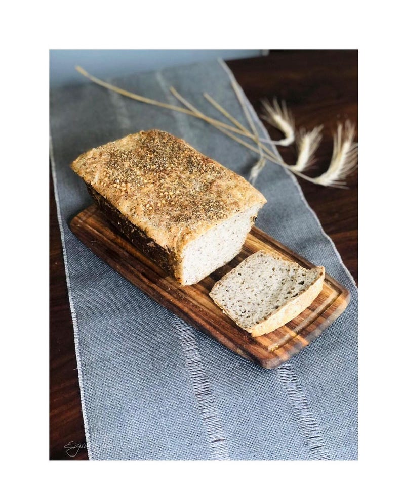 Pode incluir: Um p&atilde;o artesanal retangular com uma crosta dourada coberta de sementes e ervas, sobre uma t&aacute;bua de corte de madeira. Uma fatia est&aacute; cortada e colocada ao lado do p&atilde;o, revelando um miolo interior claro e poroso.