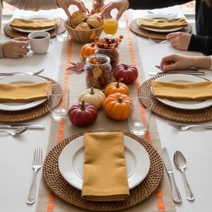 May include: A table setting featuring a burlap table runner with orange stitched borders, white plates, mustard yellow napkins, and woven placemats. The centerpiece includes small pumpkins, glass jars with berries, and a basket of bread rolls.