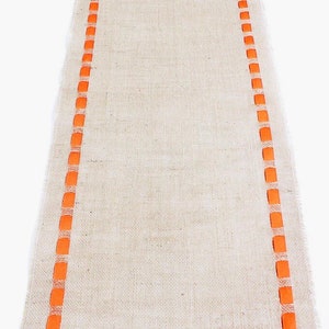 May include: A long, rectangular table runner made of natural tan burlap fabric. The runner features two parallel vertical borders consisting of a woven orange ribbon pattern that runs along both sides of the length of the textured material.