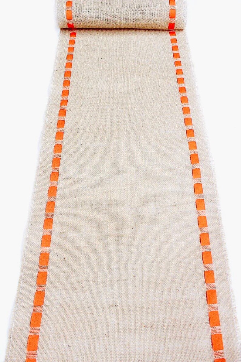May include: A long, rectangular table runner made of natural tan burlap fabric. The runner features two parallel vertical borders consisting of a woven orange ribbon pattern that runs along both sides of the length of the textured material.