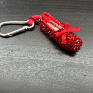 May include: A miniature red ballet slipper keychain, encrusted with sparkling red rhinestones. The slipper has a small red bow and is attached to a silver-colored metal clip with a red fabric loop.