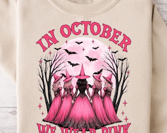 May include: A beige sweatshirt with a graphic that reads "IN OCTOBER WE WEAR PINK." The graphic features a pink moon, bats, trees, and five witches in pink dresses and hats. A Halloween-themed design.