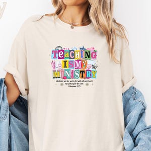 May include: An ivory short-sleeve t-shirt featuring a colorful graphic print with the text "TEACHING IS MY MINISTRY" and "Whatever you do, work at it with all your heart, as working for the Lord, Colossians 3:23". The shirt is layered with a blue denim jacket.
