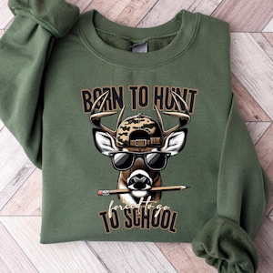 May include: An olive green sweatshirt with the text "Born to Hunt, Forced to go to School." The graphic features a deer wearing sunglasses and a camouflage hat, holding a pencil. The sweatshirt is folded on a light wood-grain surface.