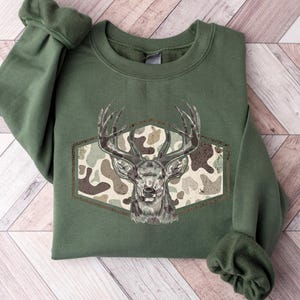 May include: An olive green sweatshirt featuring a detailed illustration of a deer head with large antlers. The deer is set against a camouflage pattern within a brown hexagonal frame. The sweatshirt has cuffed sleeves and a crew neck.