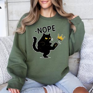 May include: Green sweatshirt featuring a black cat kicking a gold crown. The word "NOPE" is above the cat. The cat has yellow eyes and a grumpy expression. The sweatshirt is a solid colour.