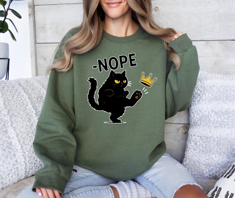 May include: Green sweatshirt featuring a black cat kicking a gold crown. The word "NOPE" is above the cat. The cat has yellow eyes and a grumpy expression. The sweatshirt is a solid colour.
