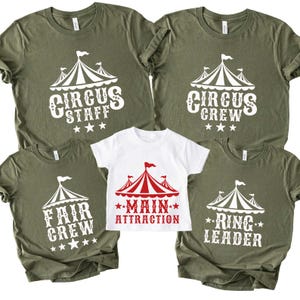 May include: Olive green and white t-shirts with circus-themed designs. The shirts feature text like "Circus Staff," "Circus Crew," "Fair Crew," and "Ring Leader." A white shirt displays "Main Attraction" in red.