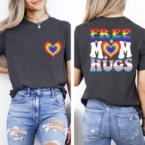 May include: A dark gray t-shirt featuring a small rainbow heart graphic on the front left chest. The back of the shirt displays the text FREE MOM HUGS in colorful lettering, with a rainbow heart, small white flowers, and sparkles.