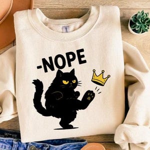 May include: Cream-colored sweatshirt with a black cat graphic kicking a gold crown. The word "-NOPE" is printed above the cat. The cat has yellow eyes and a grumpy expression. A fun, casual design.