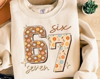 May include: A cream-colored sweatshirt with the numbers "67" in a floral pattern. The word "six" is above the numbers, and "seven" is below. The design includes star accents.