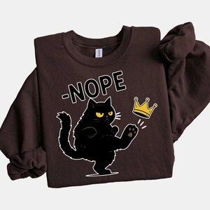 May include: A dark brown sweatshirt with a black cat illustration kicking a gold crown. The word "-NOPE" is above the cat in white block letters. The sweatshirt is a casual style.