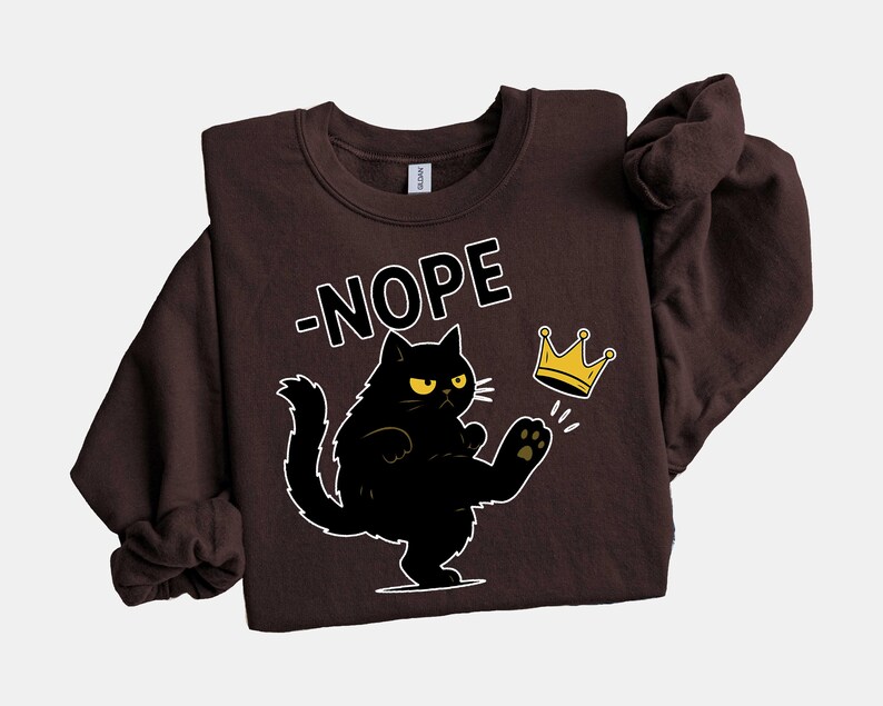 May include: A dark brown sweatshirt with a black cat illustration kicking a gold crown. The word "-NOPE" is above the cat in white block letters. The sweatshirt is a casual style.