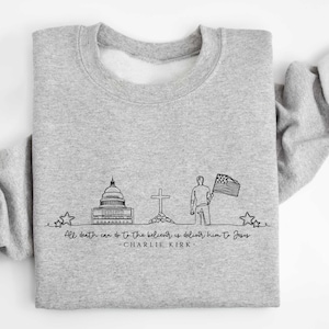 May include: A light gray sweatshirt with a black line drawing of the U.S. Capitol building, a cross, a person holding a flag, and the text "All death can do to the believer is deliver him to Jesus - Charlie Kirk".