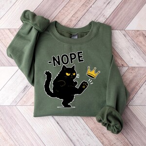 May include: An olive green sweatshirt featuring the word "-NOPE" above a black cat illustration kicking a gold crown. The cat has yellow eyes and white whiskers. The sweatshirt is folded on a light brown and white wooden surface.