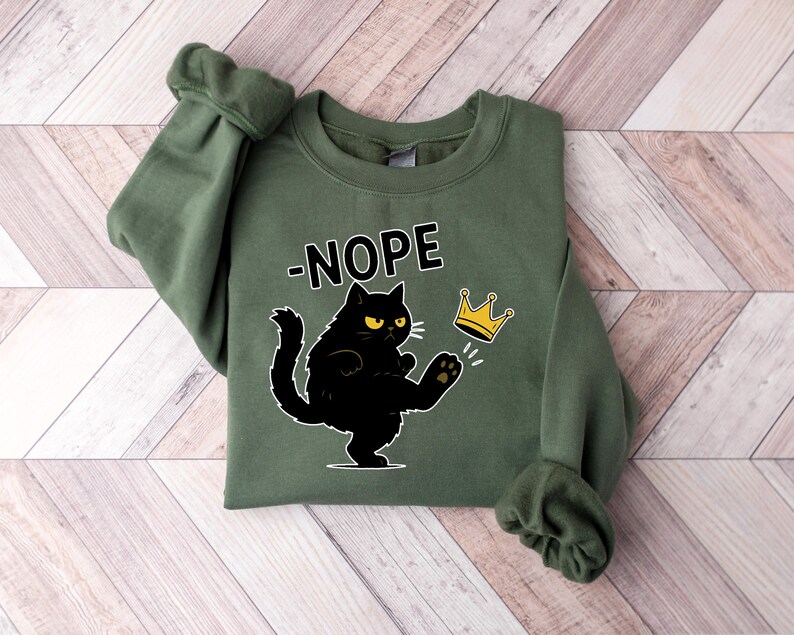 May include: An olive green sweatshirt featuring the word "-NOPE" above a black cat illustration kicking a gold crown. The cat has yellow eyes and white whiskers. The sweatshirt is folded on a light brown and white wooden surface.