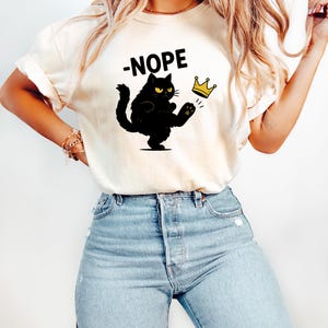 May include: A cream-coloured t-shirt with a black cat kicking a gold crown, and the word "-NOPE" above. The cat has yellow eyes and is in a defiant pose. The shirt is worn with light blue jeans.