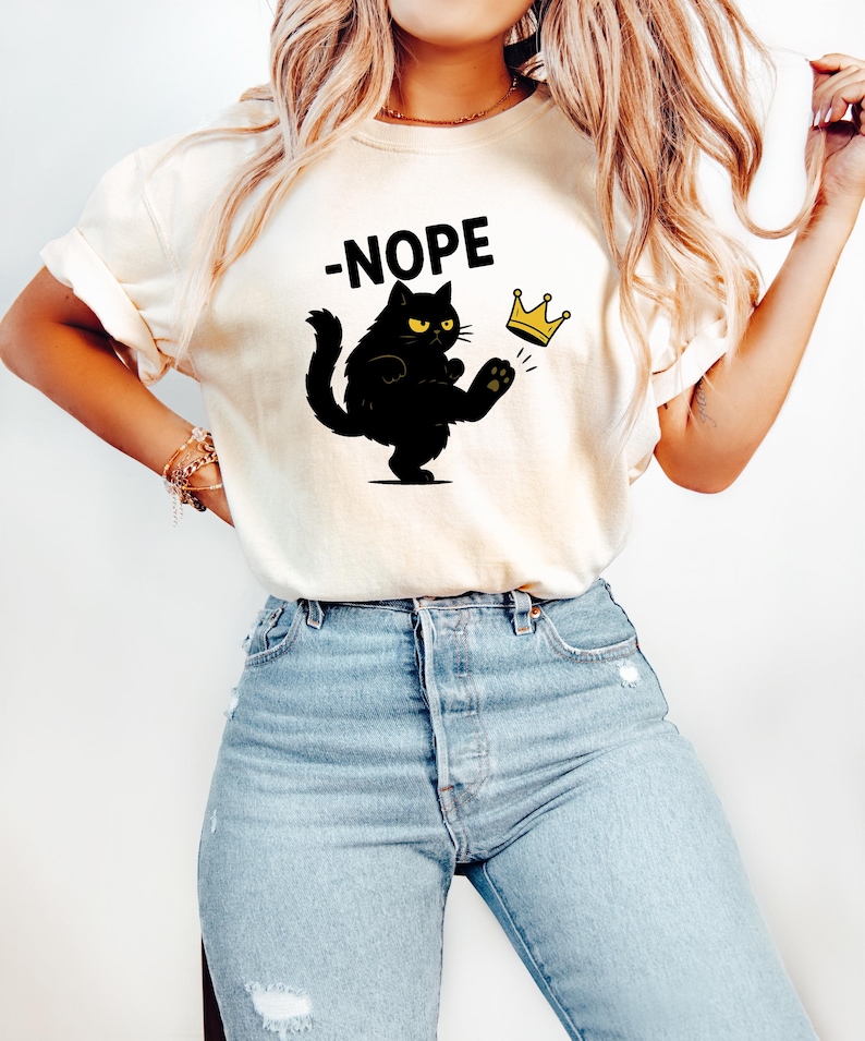 May include: A cream-coloured t-shirt with a black cat kicking a gold crown, and the word "-NOPE" above. The cat has yellow eyes and is in a defiant pose. The shirt is worn with light blue jeans.