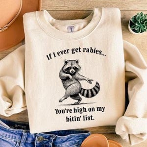 May include: Cream-colored sweatshirt with a raccoon graphic and the text "If I ever get rabies... You're high on my bitin' list." The raccoon is depicted in a dancing pose. The sweatshirt is made of a soft material.
