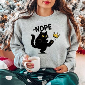 May include: A light grey sweatshirt featuring the word "-NOPE" above a black cat kicking a gold crown. The cat has yellow eyes. The sweatshirt is part of a casual outfit.