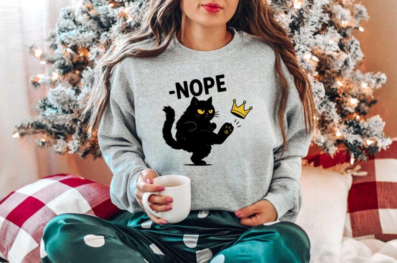 May include: A light grey sweatshirt featuring the word "-NOPE" above a black cat kicking a gold crown. The cat has yellow eyes. The sweatshirt is part of a casual outfit.