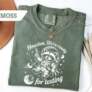 May include: Moss green t-shirt with a white graphic of a raccoon in a spacesuit, the text "Houston, We're ready for testing", and a gold necklace. The shirt is made of a soft material.