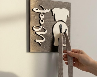 May include: Wooden dog leash holder with a dark gray background. The design features a dog's silhouette and the word "Woof" in white script. Two hooks hold a leash. A hand is holding the leash.