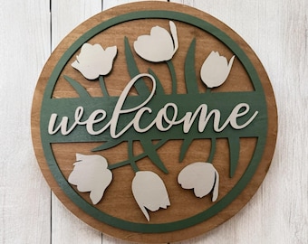 May include: A round wooden welcome sign with a brown base, green border, and white tulips. The word "welcome" is cut out in white script across the center. The sign is hanging on a white wooden background.