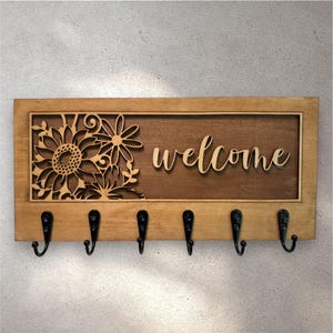 May include: Wooden welcome sign with a sunflower design and six black hooks. The sign features the word "welcome" in a cursive font. The sunflower is intricately cut out of the wood, adding a decorative touch to the entryway decor.