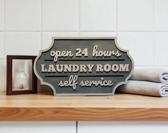 May include: A wooden sign with the words "open 24 hours LAUNDRY ROOM self service" in white lettering. The sign is gray with a decorative border. A small framed hourglass and folded towels are also visible.