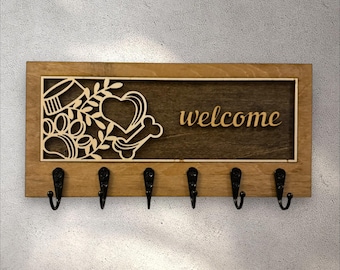 May include: Wooden key holder with a rectangular frame and six black hooks. The center features a decorative cutout with a paw print, heart, and bone design, with the word "welcome" in a script font.