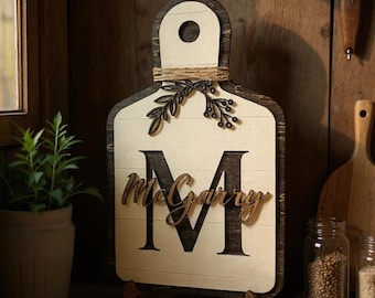 May include: Wooden decorative cutting board with a handle, featuring a large letter "M" and the name "McGarry" in a script font. The board is cream-colored with dark brown edges and a decorative twine and leaf accent.