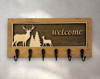 May include: Wooden key holder with a rectangular frame. The design features a deer, a fawn, and pine trees, with the word "welcome" in a script font. Six black metal hooks are attached below the design.