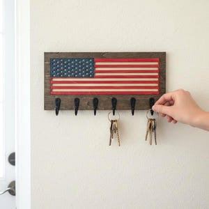 May include: Wooden key holder with an American flag design. The key holder has a dark brown frame, with red and white stripes and a blue field of stars. Six black hooks are mounted below the flag for hanging keys.
