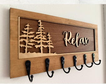 May include: Wooden wall-mounted key holder with a light brown frame and a dark brown center. The center features a laser-cut forest scene and the word "Relax" in cursive. Six black metal hooks are attached below.