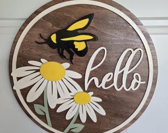 May include: Round wooden sign with a dark brown background, a white border, and the word "hello" in cursive. The sign features a yellow and black bee and two white daisies with yellow centers and green stems.